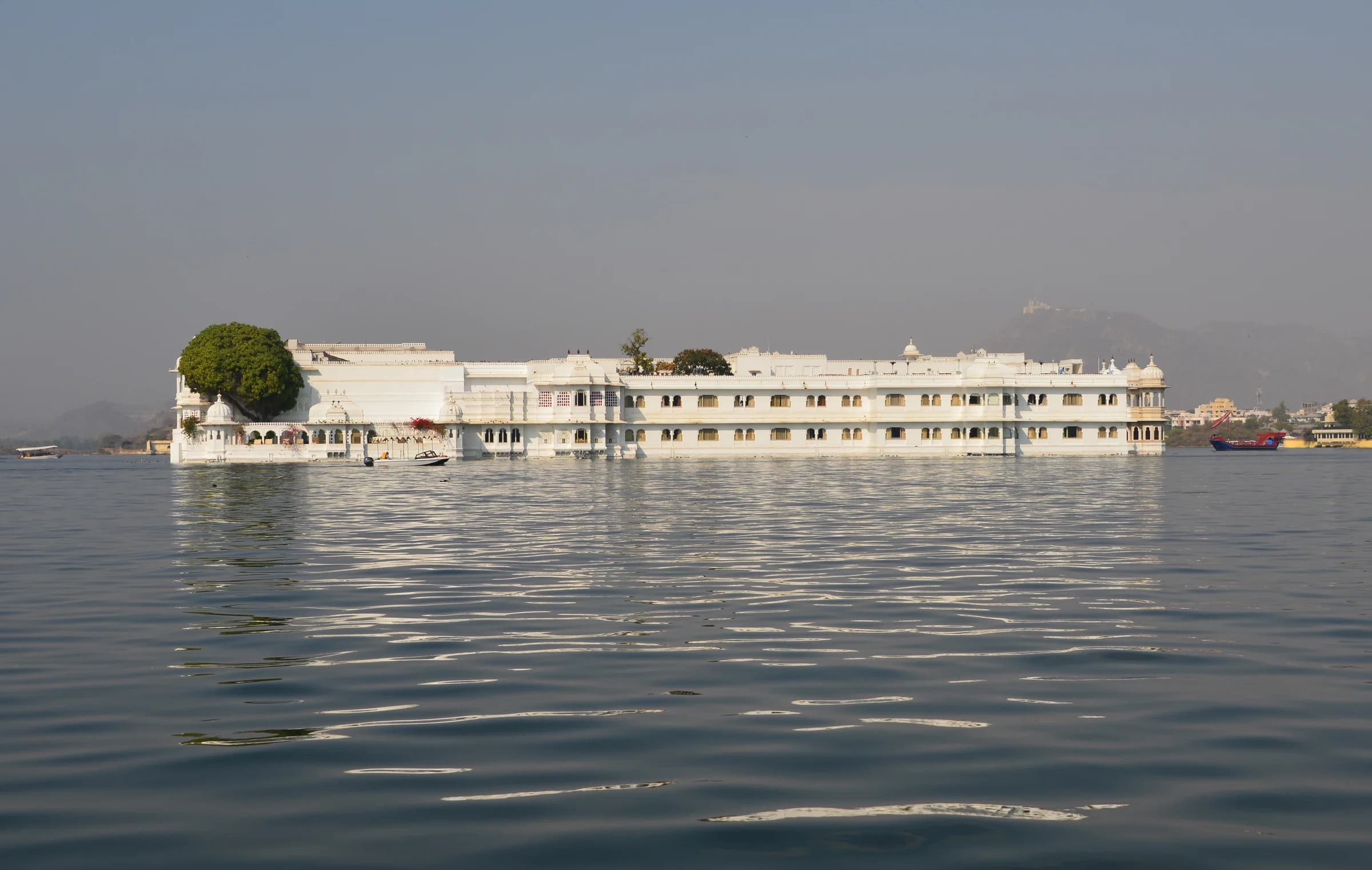Taj Lake Palace viewed from across Lake Pichola at sunset — the white marble palace floating on the glassy water, the Aravalli Hills softening into silhouette, the entire scene suspended between sky and reflection