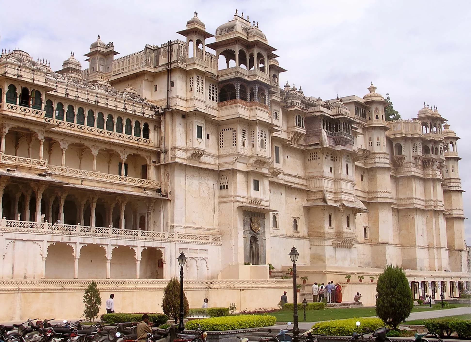 City Palace Udaipur viewed from Lake Pichola at sunset — the entire eastern shore dominated by the sprawling complex, a cascade of domes, cupolas, balconies, and towers rising from the water's edge to the hilltop, every surface glowing gold in the evening light