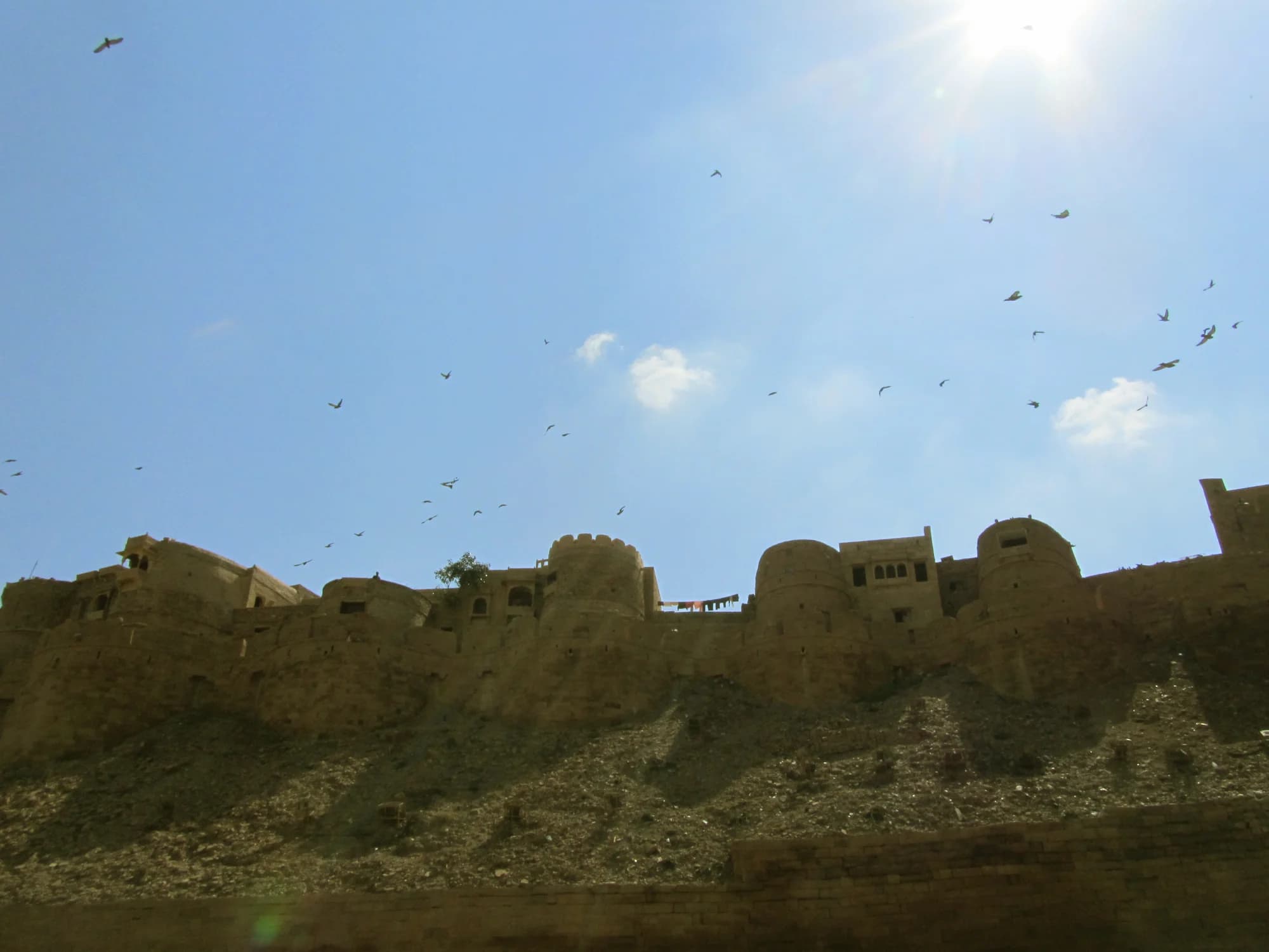 Suryagarh at sunrise — the golden sandstone fortress emerging from the Thar Desert haze, its crenellations and domed chhatris silhouetted against a sky of pale rose and amber, the surrounding dunes empty of any sign of human presence