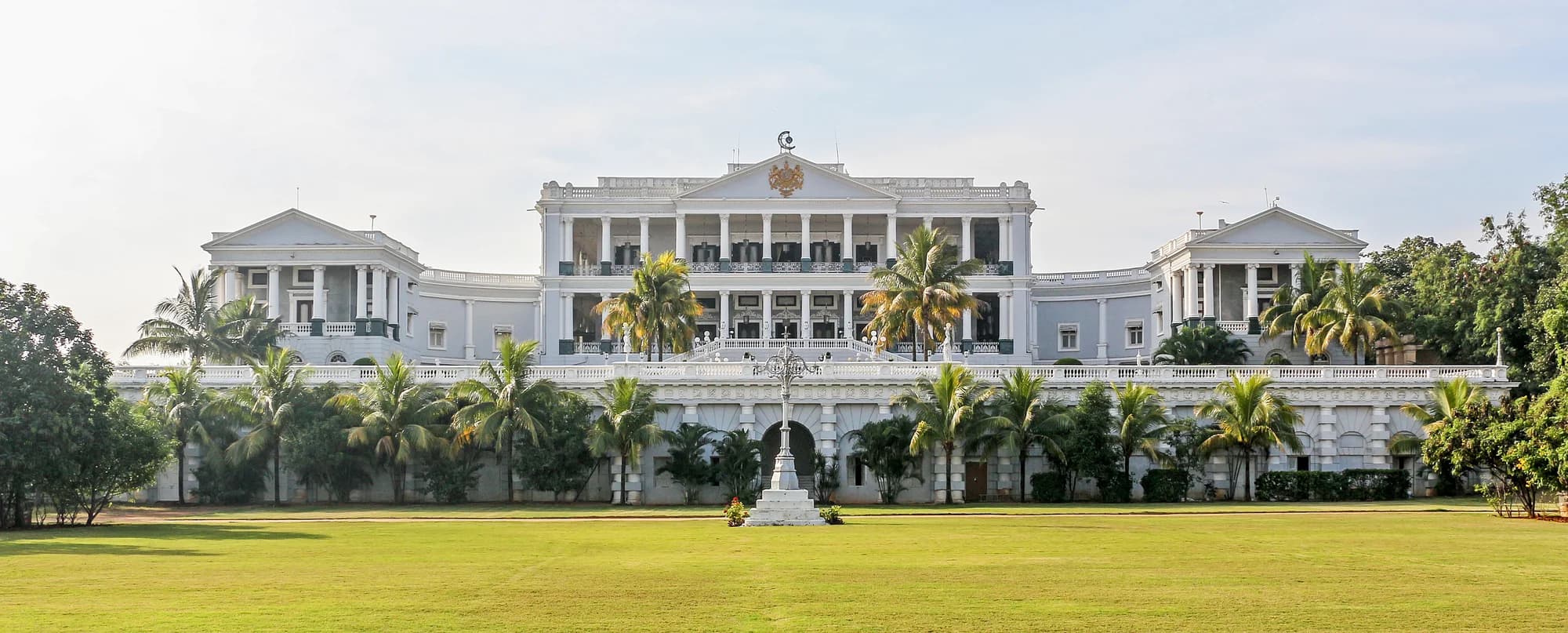 Falaknuma Palace at twilight — the white marble facade glowing rose-gold against the Deccan sky, the city of Hyderabad sprawled two thousand feet below, the palace's distinctive dome and turrets silhouetted against gathering dusk