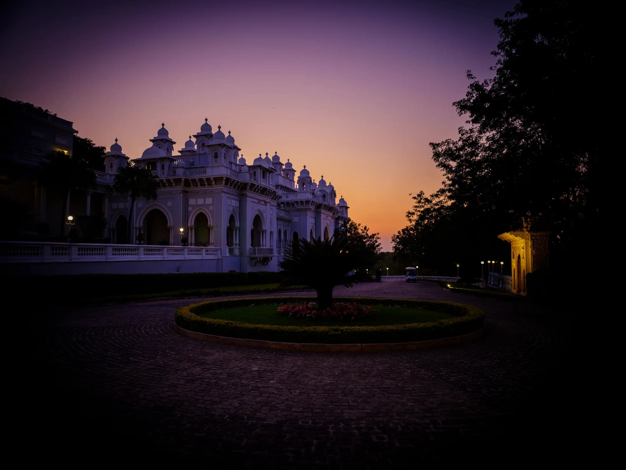 Falaknuma Palace at blue hour — the white marble facade illuminated from within, the crescent wings embracing the formal gardens, Hyderabad's city lights glittering two thousand feet below, the dome catching the last amber light of the Deccan sunset
