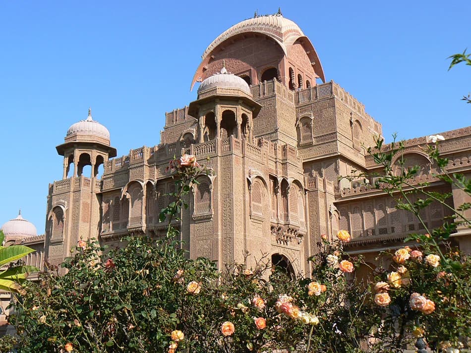The inner courtyard at midday — carved columns casting geometric shadows across marble floors, a central fountain playing softly, the red sandstone walls glowing in the fierce desert light filtered through centuries-old jali screens