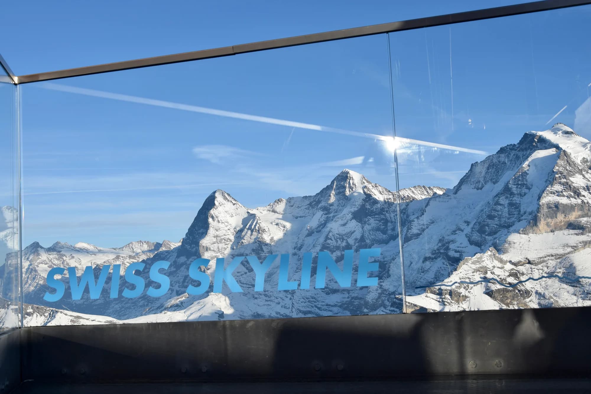 Paragliders descending over Interlaken — the brightly coloured canopies silhouetted against the blue of Lake Thun, the Jungfrau massif filling the southern sky, the town reduced to a miniature of hotels, churches, and the green ribbon of the Aare River connecting the two lakes