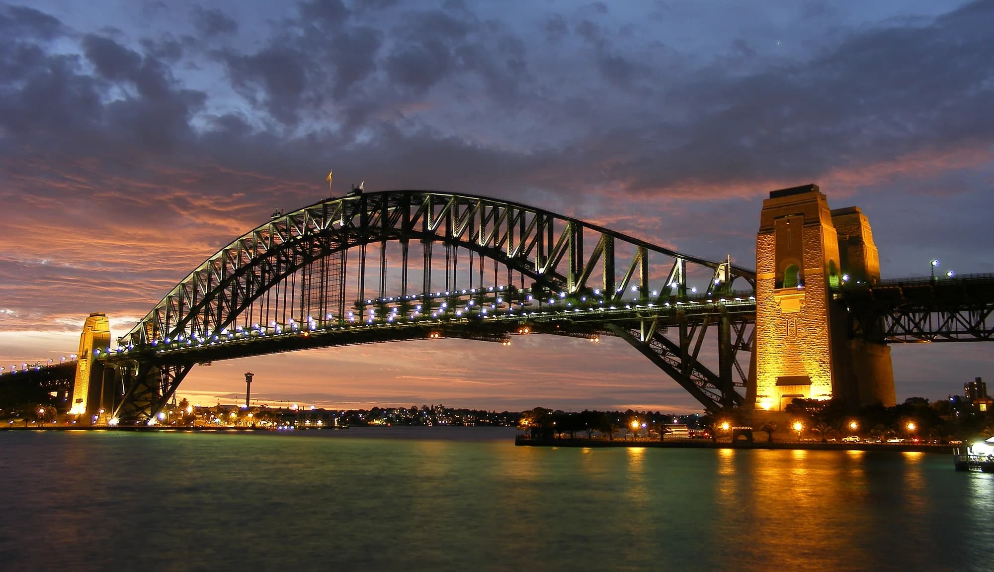 Climbers on the upper arch of the Sydney Harbour Bridge at sunrise — the Opera House below, the CBD towers catching the golden light, the Pacific Ocean stretching to the eastern horizon, the climbers' grey suits silhouetted against the steel latticework